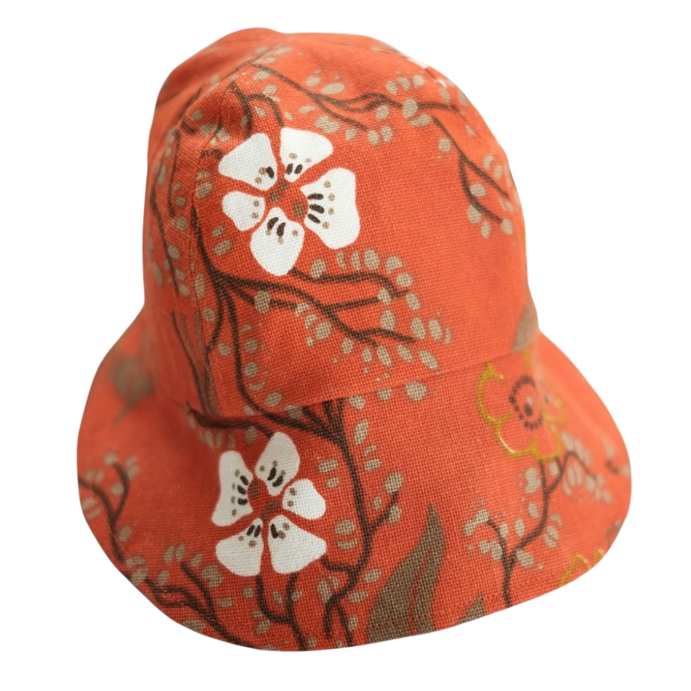 Handgjord solkeps i återbrukat orange tyg med stora vita blommor och kvistar. Fotograferad mot svart bakgrund.