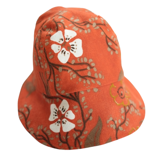 Handgjord solkeps i återbrukat orange tyg med stora vita blommor och kvistar. Fotograferad mot svart bakgrund.